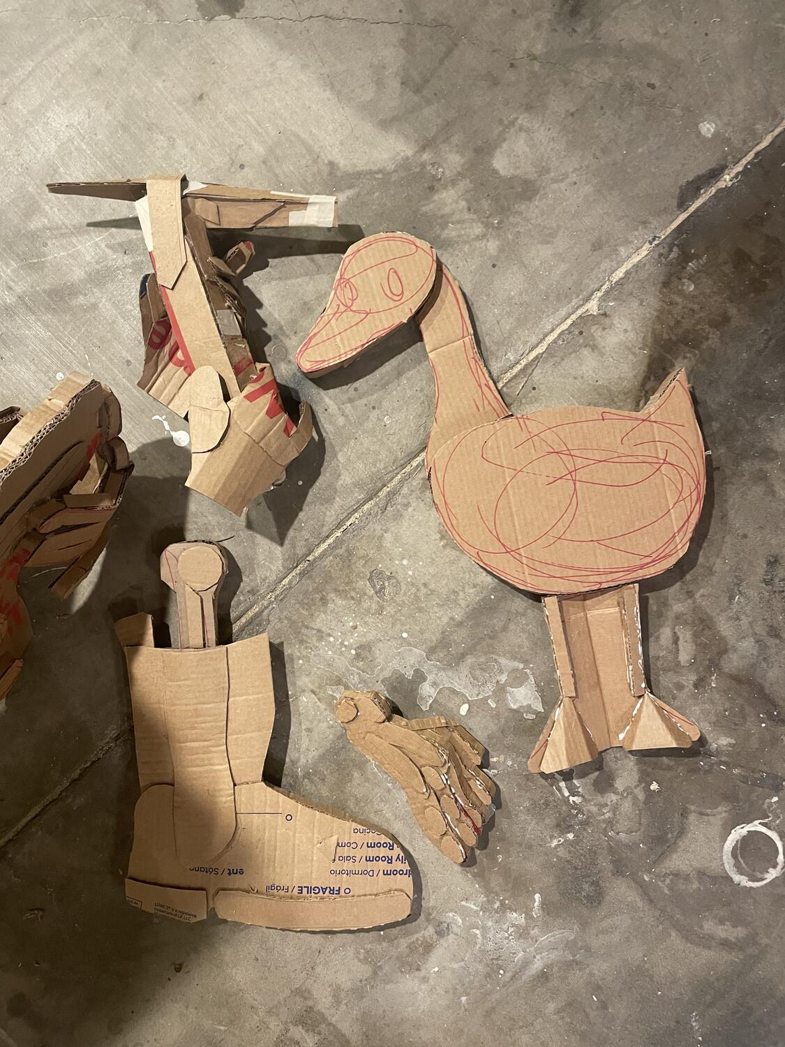 Various portions of cardboard sculpture: a duck, a boot with a lege bone sticking out, a skeletal foot, and a welding torch being held by disembodied gloves