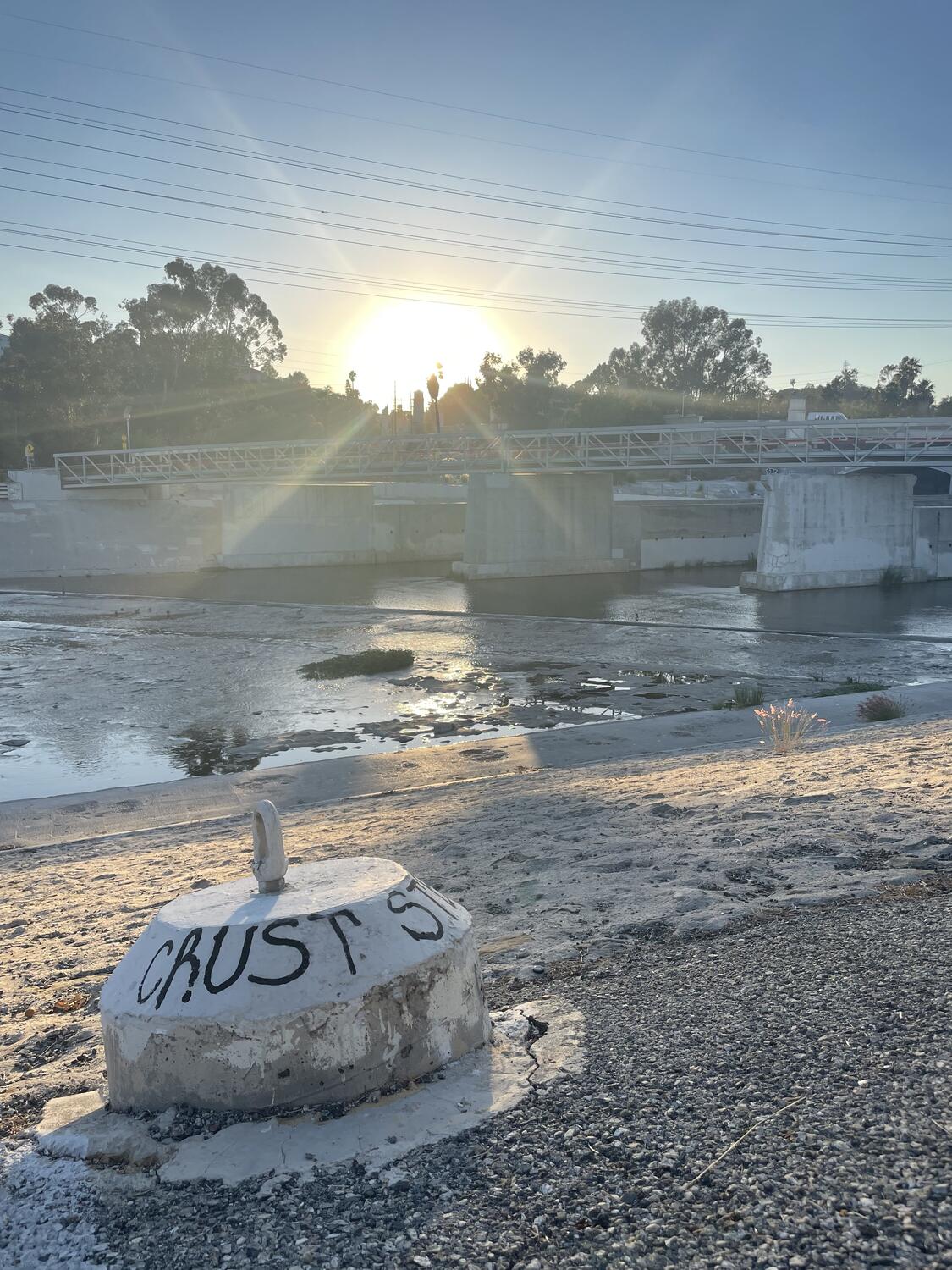 The sun setting on the LA river. The Red Car Trolley foot bridge cross in the background, a sandy portion of the bank precedes it, and in the foreground is a concrete anchor bearing a hasty graffito that reads “CRUST”