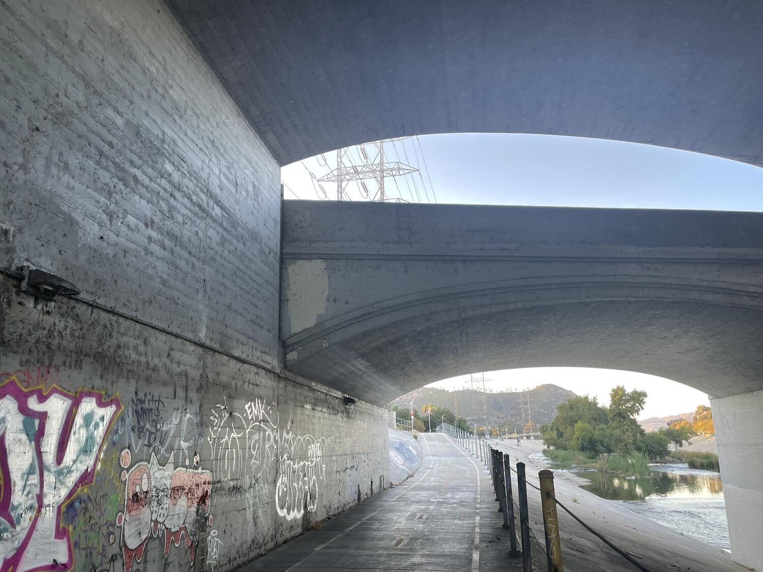 Two sections of the Glendale-Hyperion bridge, seen from the path that passes below them. They are both arched, but offset verticall, such that the upper edge of one is complete below the other and creates a little slice of sunlight. Through the gap we see power lines.