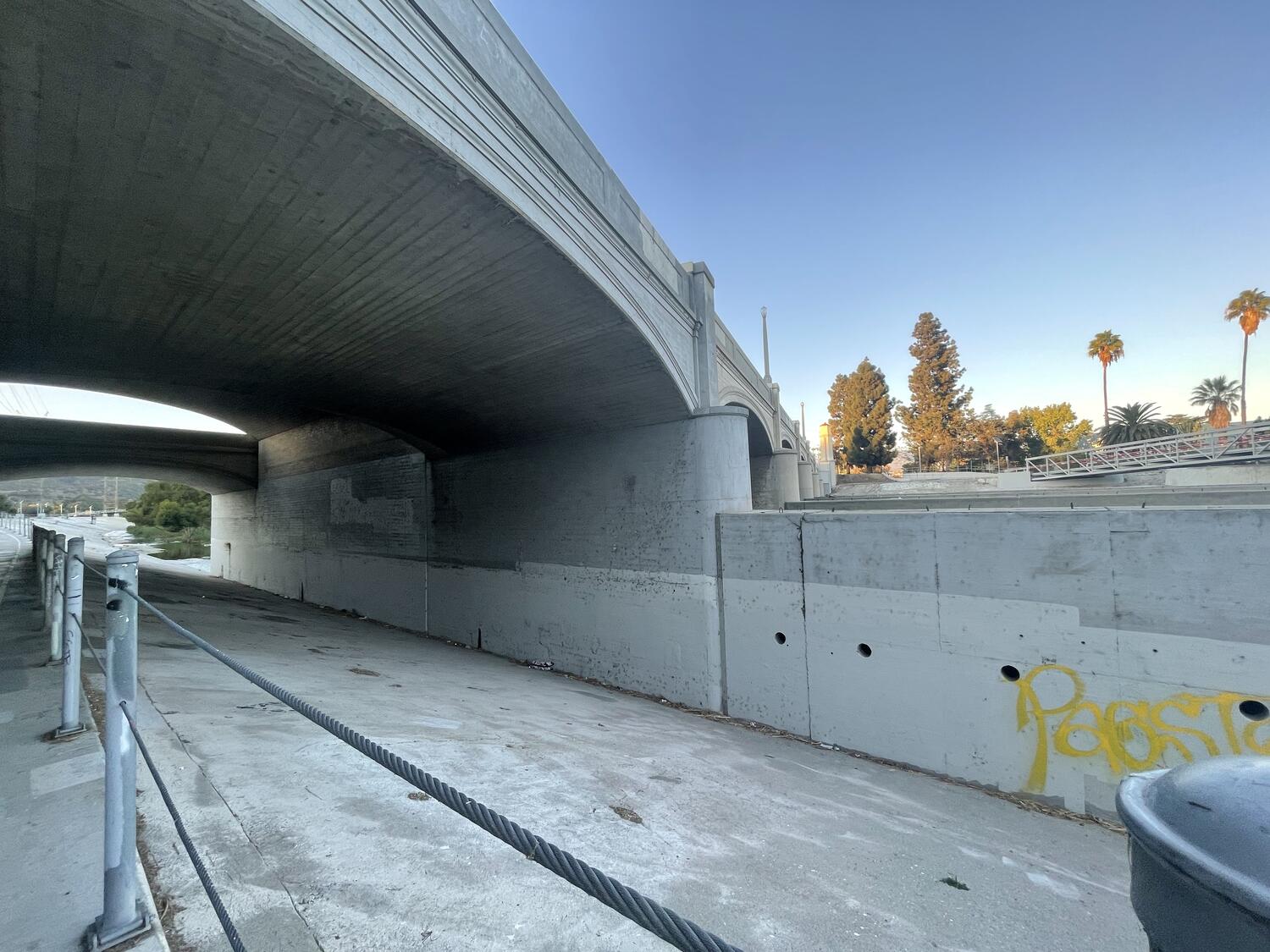 The edge of the bike path under the Glendale-Hyperion bridge. The steep slope of the bank meets with a solid wall holding up the bridge and a sharp angle, creating a strange and uninviting little angular passage