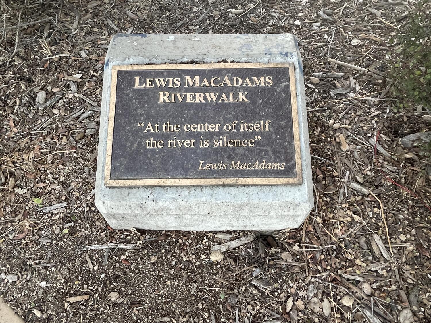 A bronze plaque mounted on a short concrete plinth, just a few inches off the ground. It reads: “Lewis MacAdams Riverwalk - At the center of itself the river is silence - Lewis MacAdams”