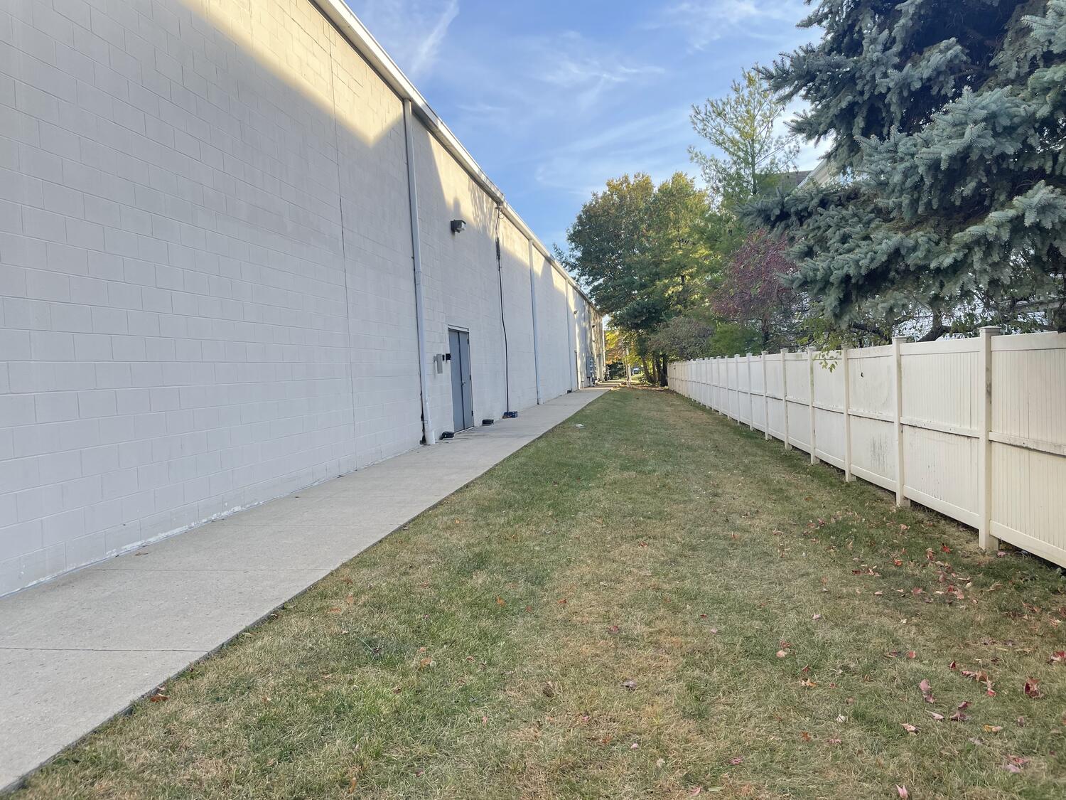 A grass alley. On one side are the plain back walls of some commercial buildings in a strip mall, and on the other is a tall white fence and trees.