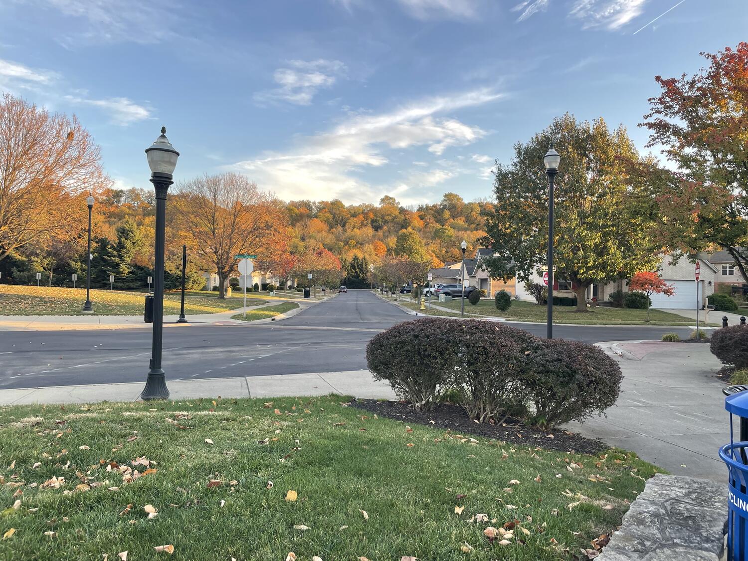 A suburban park with manicured hedges and old-fashioned lamp posts. In the center of the frame, down a short street, and throughout the whole background, there's a tall hill covered in trees, many of which are changing color for Autumn.