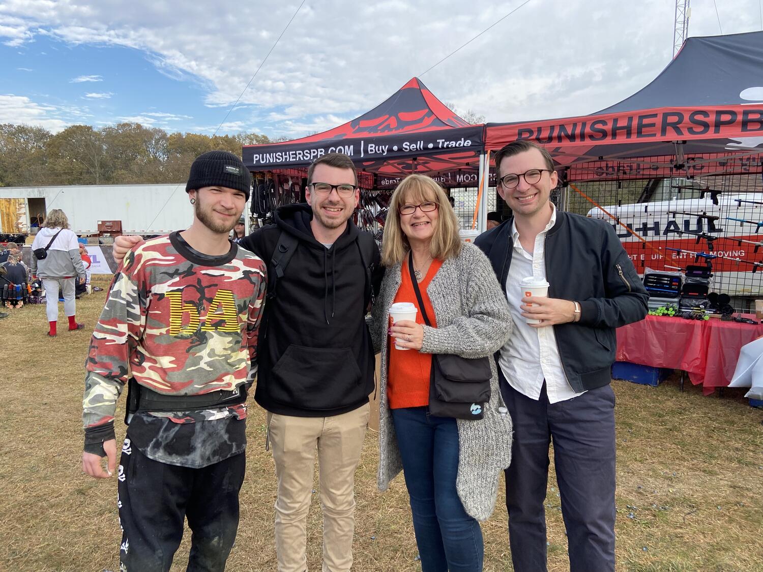 Me, my mom, and my brothers in a field. There's a tent behidn us selling paintball merchandise, and Zach is decked out in paintball gear as well.