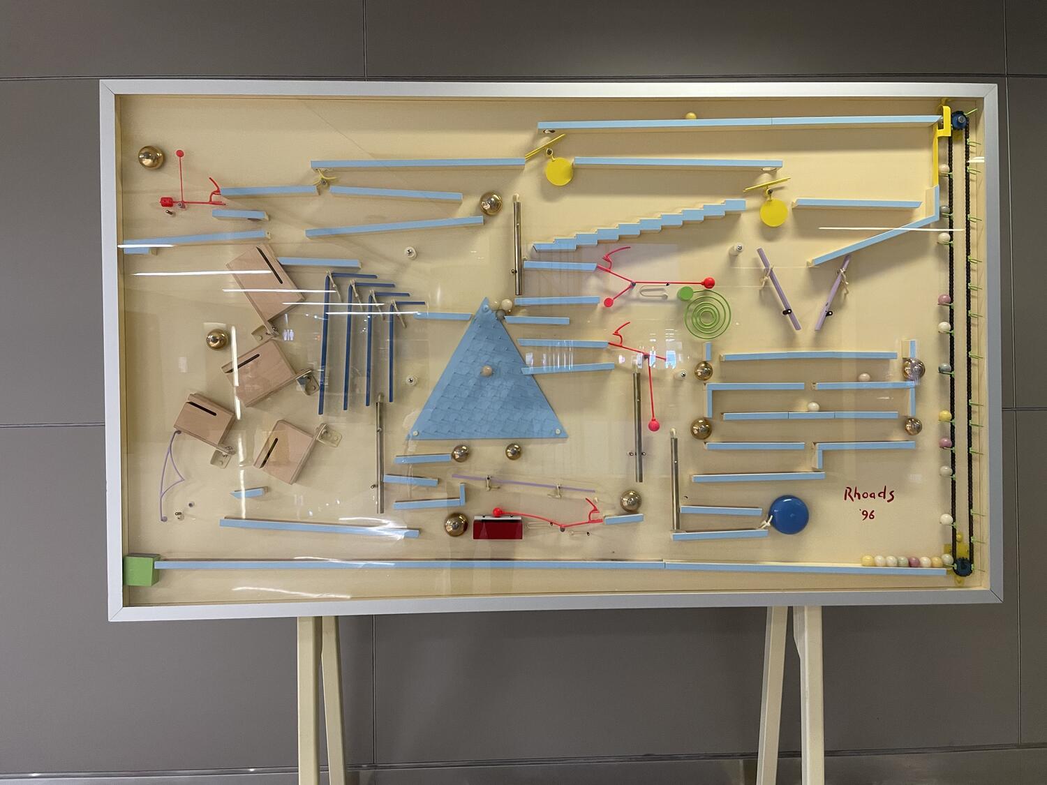 A complicated contraption of balls and ramps and bells mounted on legs near a blank wall. In the corner it's signed “Rhoads ‘96.”