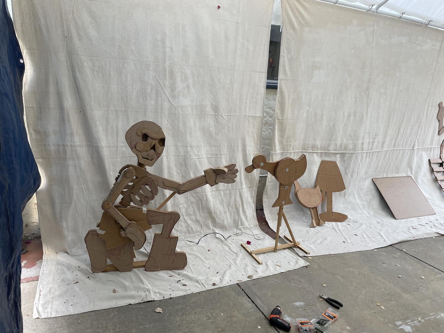 Unfinished sculptures laid out under the tent: a skeleton reaching for a duck. Some tools and hardware lie on the ground in front of them.