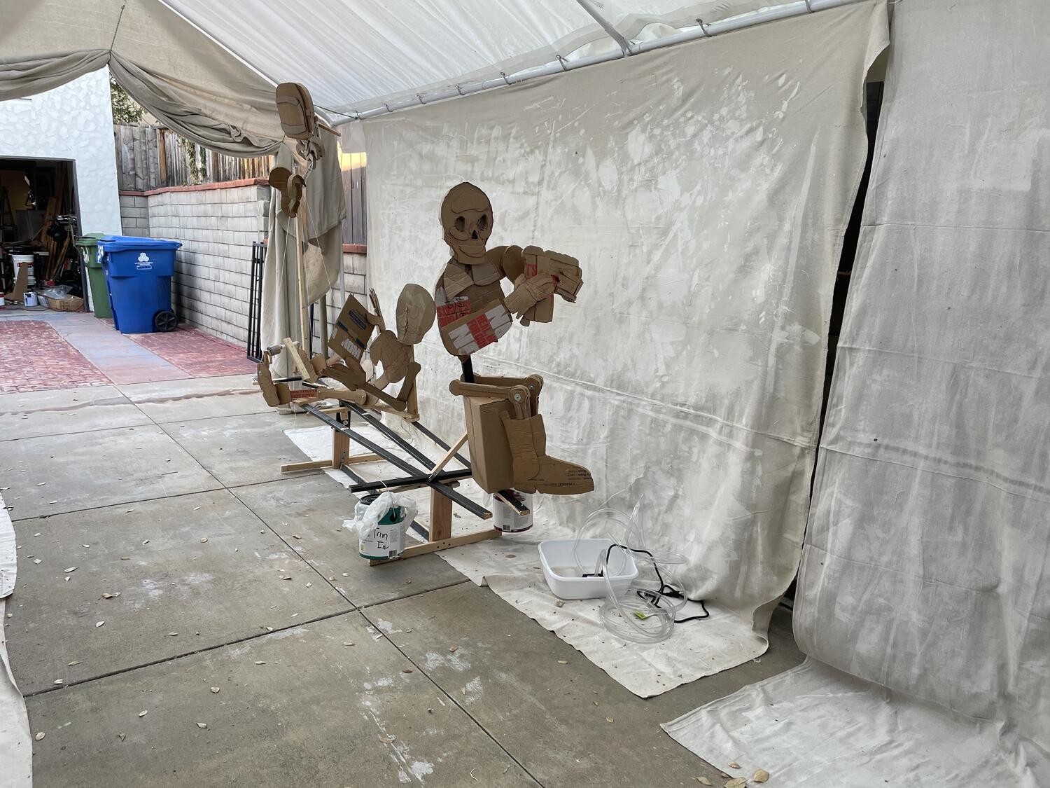 Some unfinished cardboard skeletons sitting under a tent. They're all attached to a wooden frame, and on the ground in front of them is a small plastic basin with tubes coming out of it. A messy garage is just on the edge of the frame in the background.