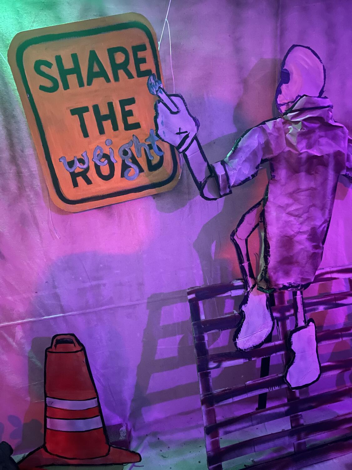 A sculpture of a skeleton in a long code, climbing up a wooden palette leaning on the wall and painting a sign. It's a road sign that originally read â€œShare The Road,â€� but it's being painted to read â€œShare The Weightâ€� instead.