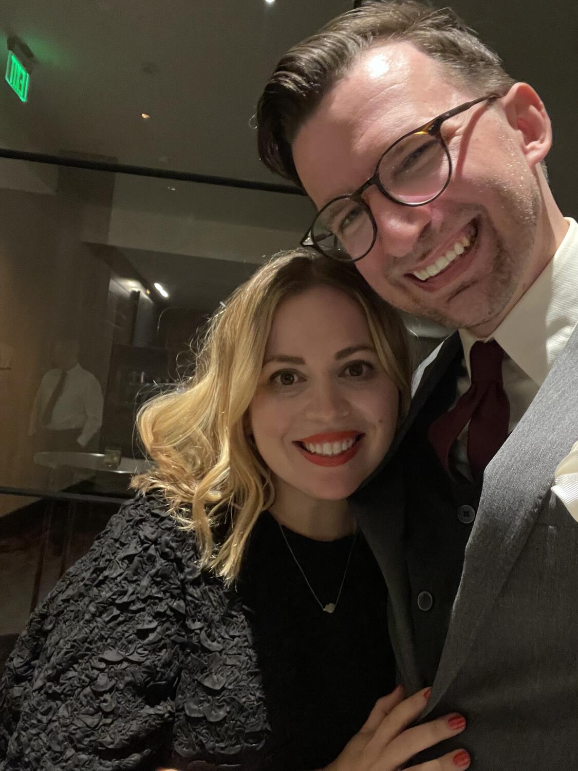 Me and Amy taking a selfie in a dark corridor of a fancy hotel. She's looking great in a black dress, I have on a grey suit and a burgundy tie and matching sweater