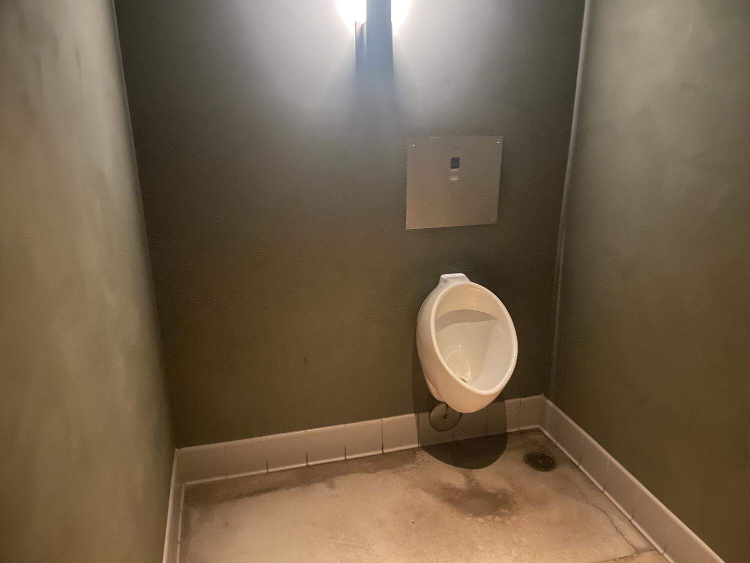 A solitary urinal in a very large restroom. It's off-center on the wall.