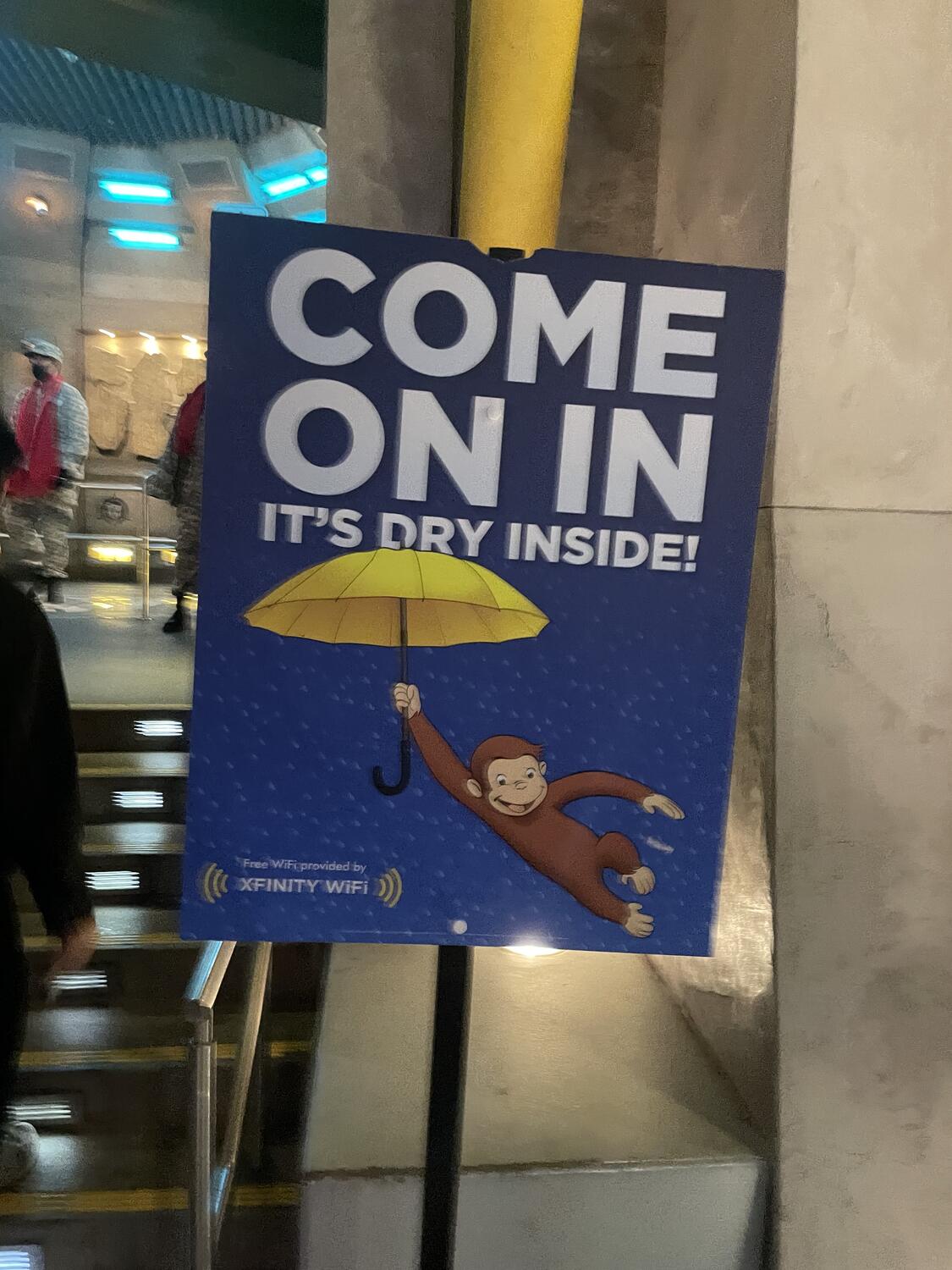 A sign standing at the entrance to Transformers: The Ride. It features Curious George with a yellow umbrella and reads "Come on in, it's dry inside!"