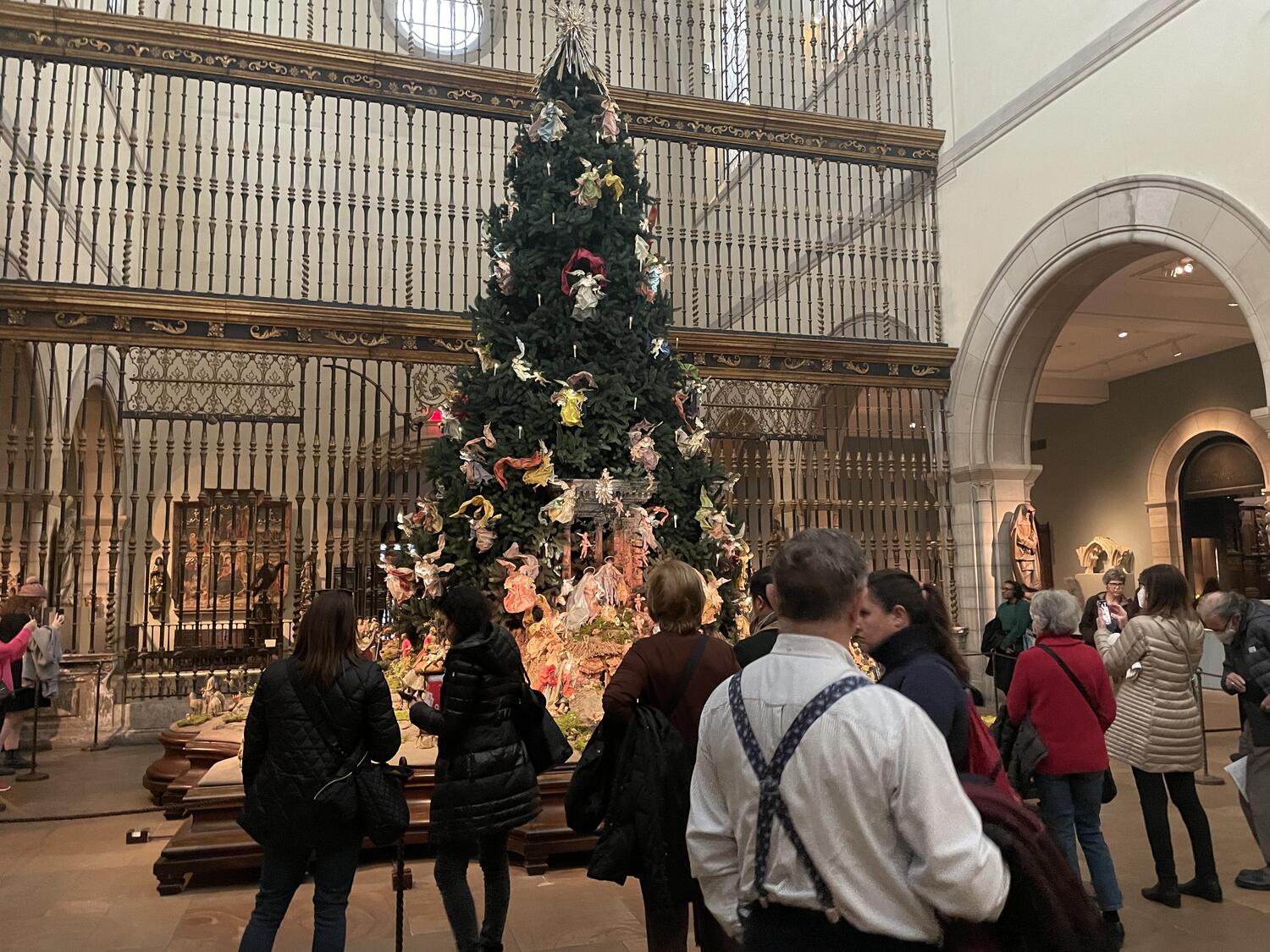 A big Christmas Tree in the Medieval Wing at the Met. It's covered in large angel sculptures and set in front of a grand metal screen that runs from floor to ceiling. Lots of people milling around.