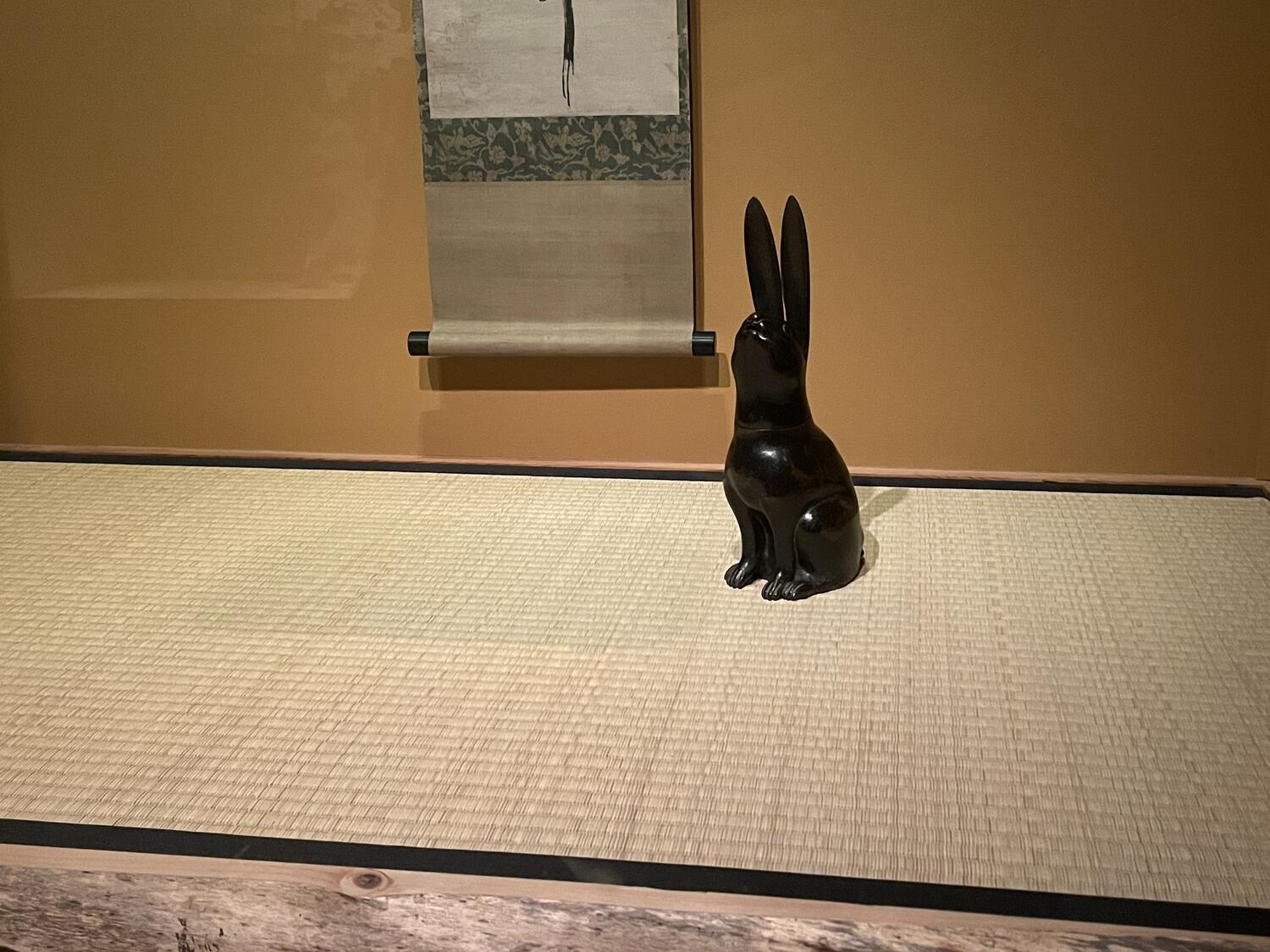 A bronze rabbit sculpture sitting on a woven mat in a display case. There's a scroll on the wall behind it bearing a minimal pattern.