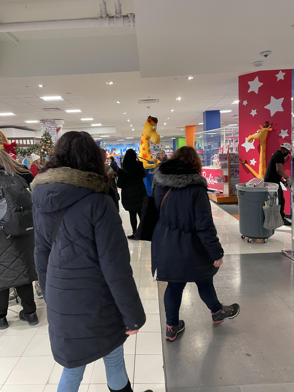 Lots of kids excitedly gathering around a giraffe mascot in the toy section of a department store