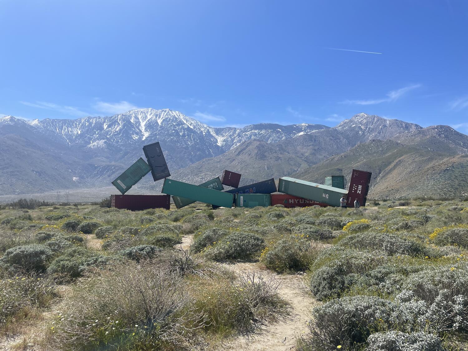 A collection of storage containers, positioned in the form of a reclining person, set in the desert against the mountains