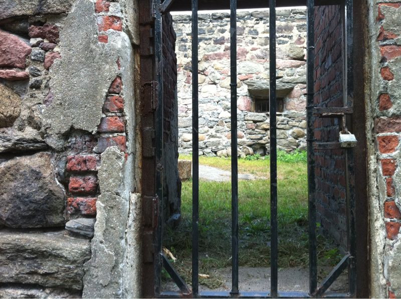 An old iron gate in a crumbling brick wall reveals the interior of a very old structure whose sunlit grass floor implies a long-missing roof