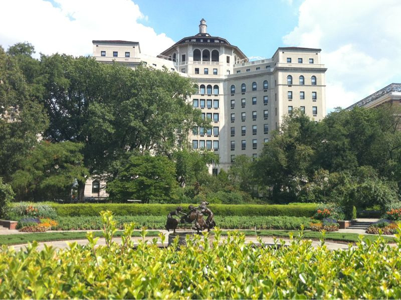 A grand building of 10 or more stories with symmetrical and imposing wings is framed by a carefully manicured garden and a bright blue sky