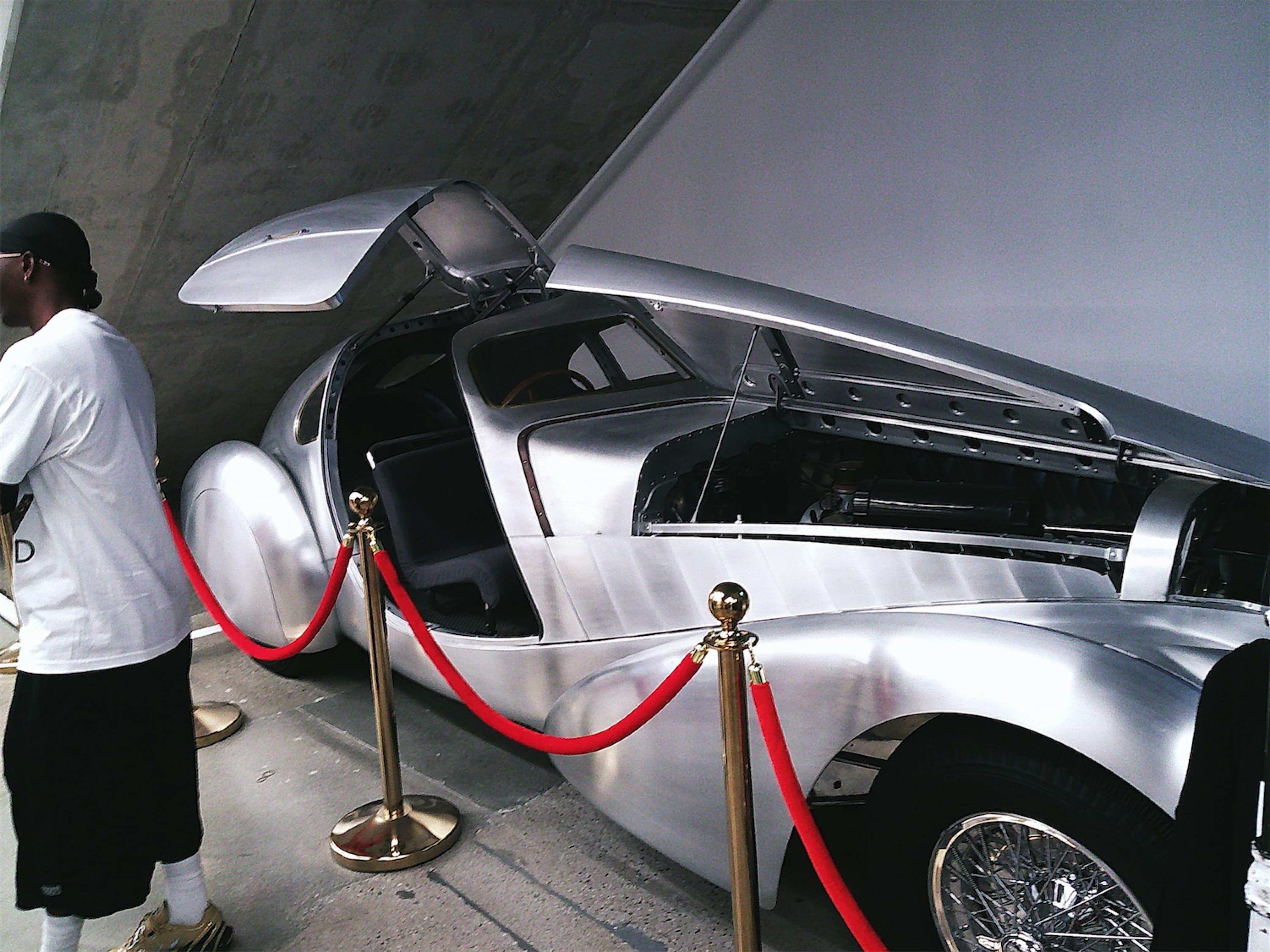 A sleek silver car behind velvet ropes, the hood and one gull-wing door aloft