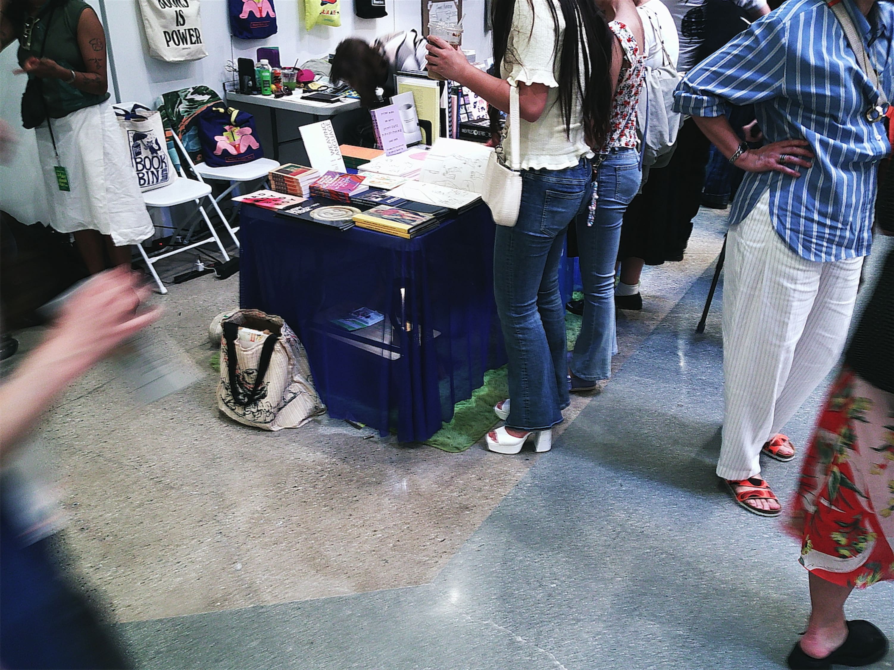A folding table covered with a sheer blue tablecloth conceals a tiny desk with a tiny lamp on a green carpet. People stand around the table, maybe unaware of the opportunities beneath it.