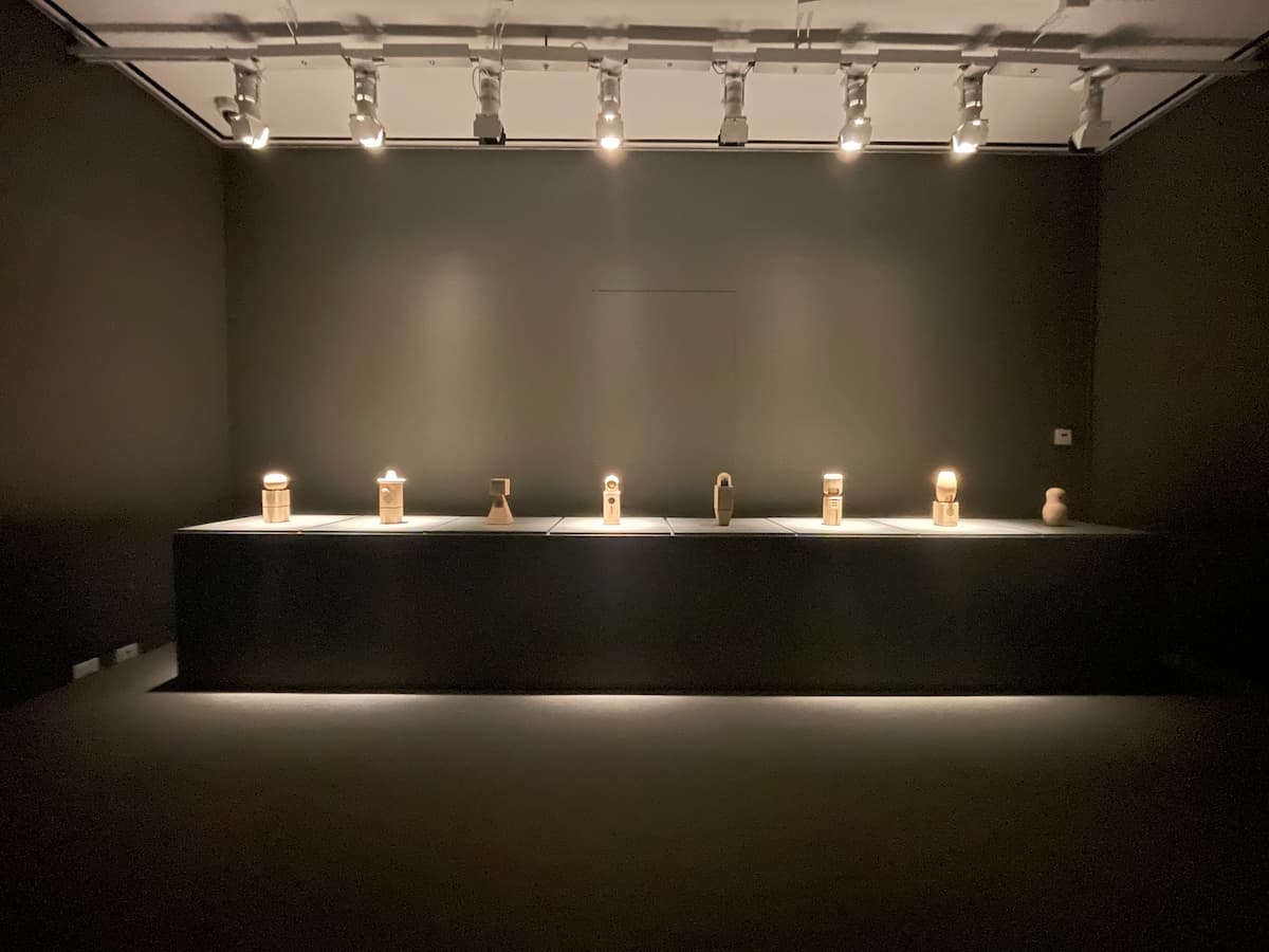 Eight wooden figurines on a wide plinth in a dark room. Four of them are dramatically lit from above; the rest are in shadow.