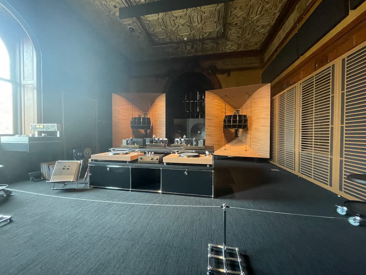 A complicated display in an old ornate room. Two huge wooden speakers, probably eight feet tall, are in the far corners. In front of them is a low table filled with turntables and difficult-to-identify boxes littered with vacuum tubes, probably?