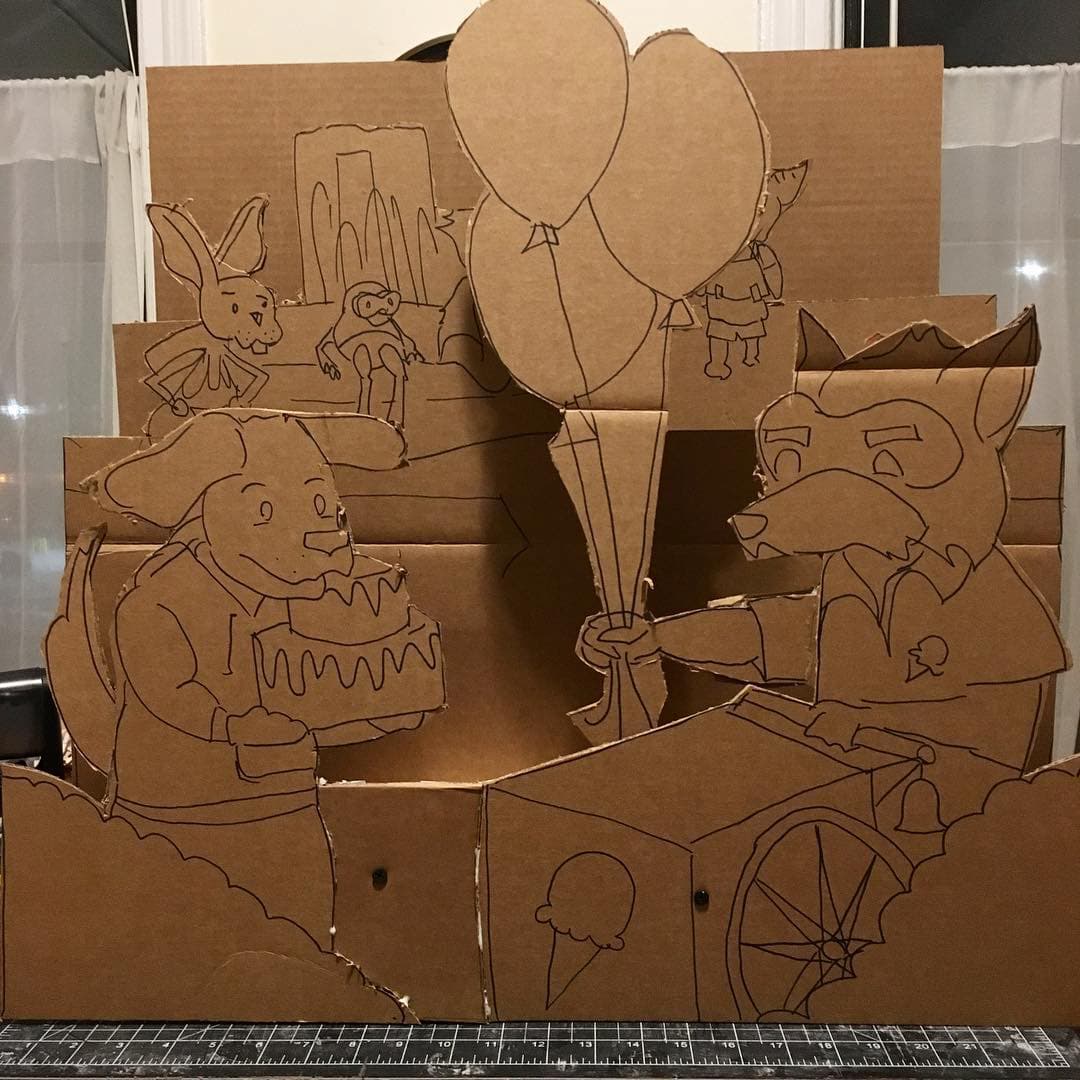 The aforementioned model of Bobby's Birthday: a few layers of cardboard cut out and stood up like a diorama, showing an anthropomorphic dog holding a big birthday cake and accepting some balloons from a raccoon.